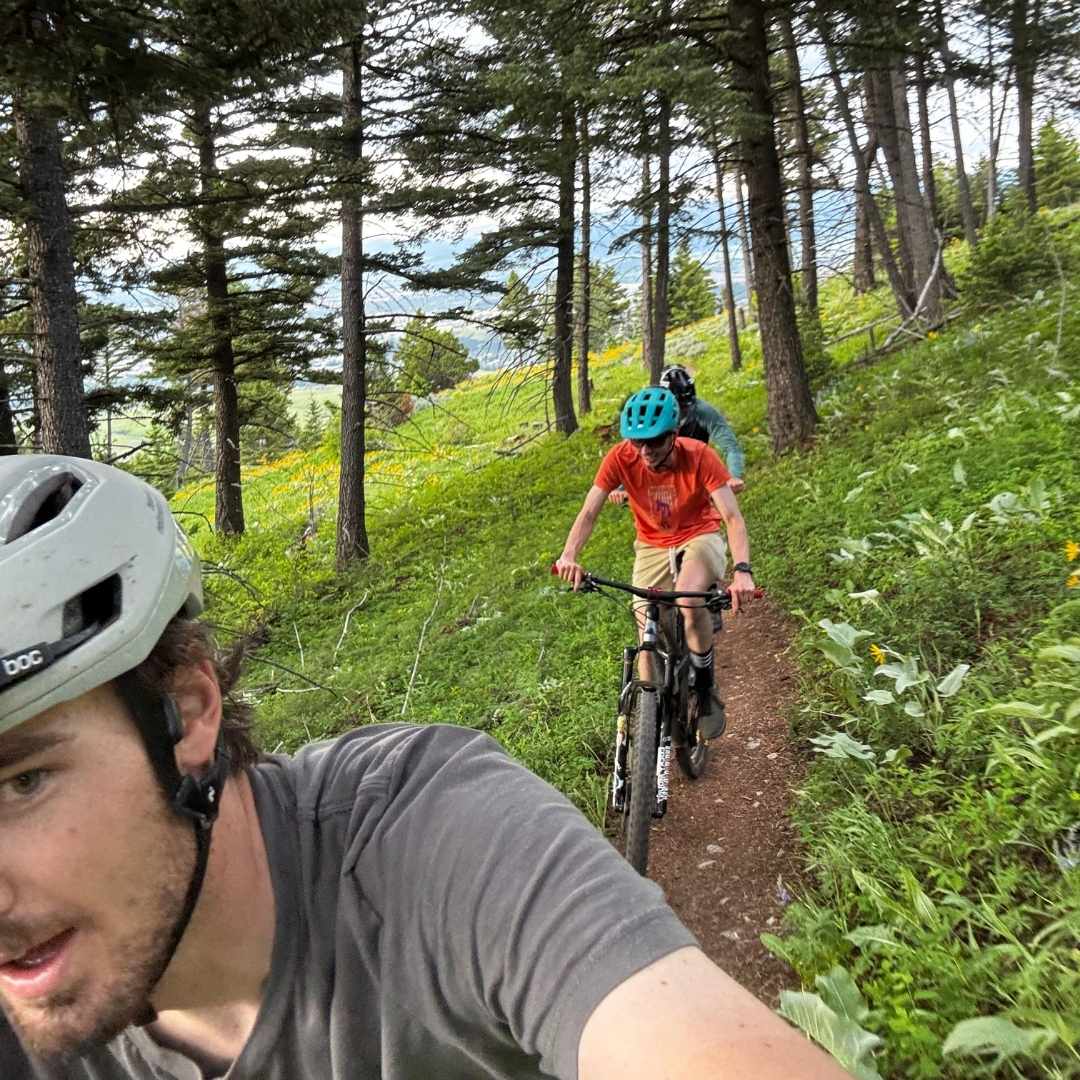 selfie shot of two mountain bikers riding through green trees on dirt trail